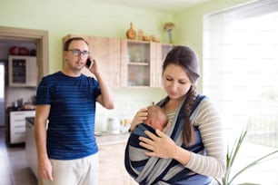 Beautiful young mother with her newborn baby son in sling at home. Father holding smart phone, making phone call.