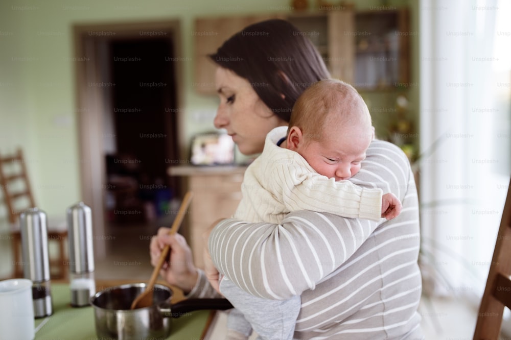 Beautiful young mother at home in the kitchen holding her newborn baby son, cooking, mixing something in pan