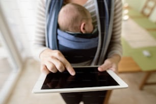Unrecognizable young mother with her newborn baby son in sling at home, holding tablet, writing something