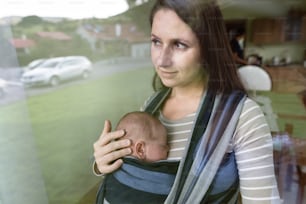 Beautiful young mother with her newborn baby son in sling at home