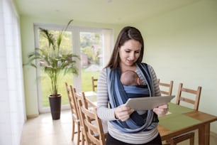 Beautiful young mother with her newborn baby son in sling at home, holding tablet, reading something