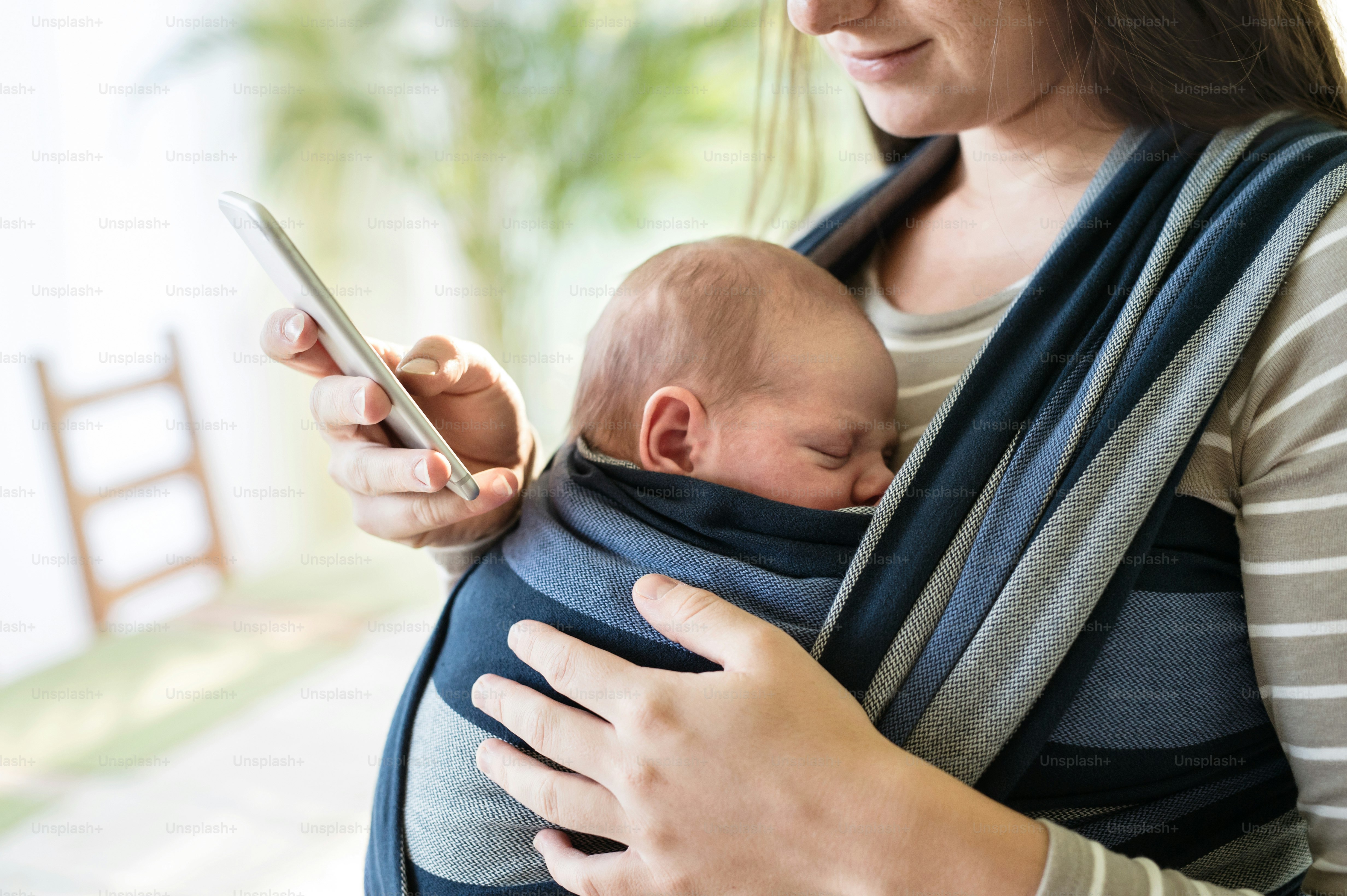 Unrecognizable young mother with her newborn baby son in sling at home, holding smart phone, texting