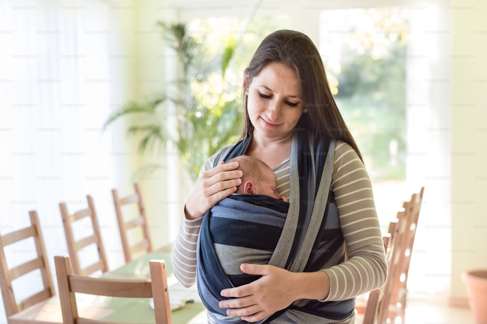 Beautiful young mother with her newborn baby son in sling at home