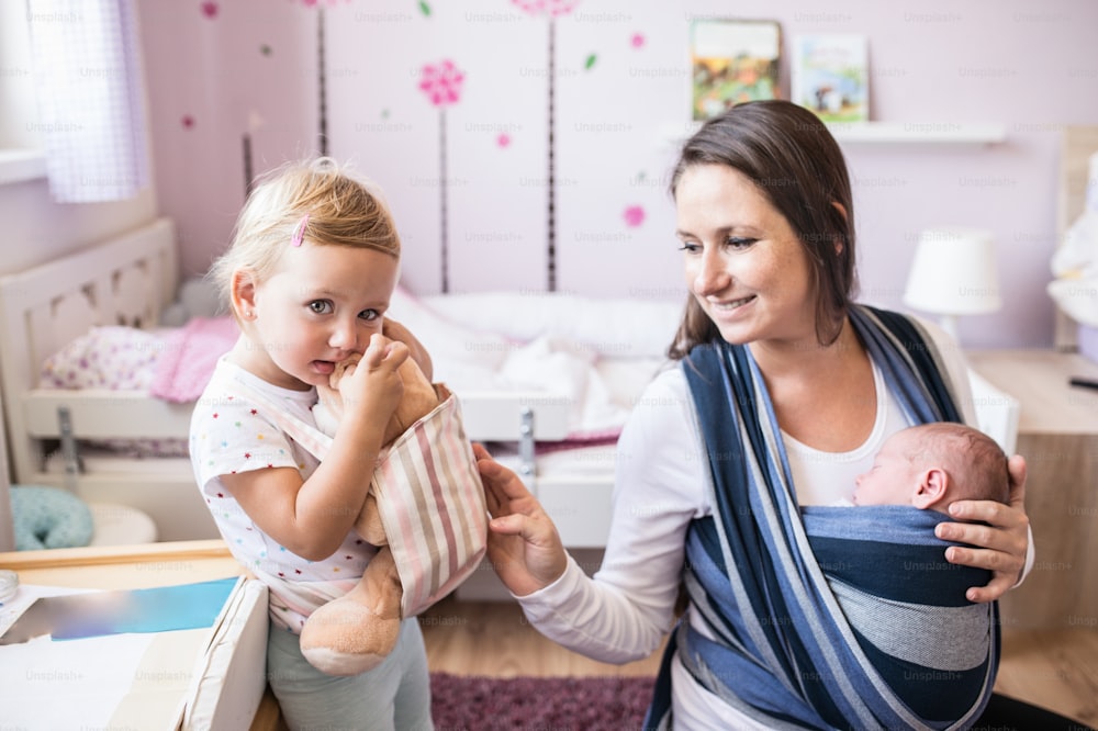 Beautiful young mother with her newborn son in sling and her daughter with teddy bear in baby carrier at home