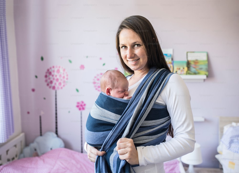 Hermosa joven madre envuelve a su hijo recién nacido en cabestrillo en su habitación