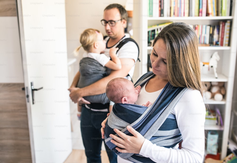 Beautiful young mother with her newborn son in sling and father with his daughter in baby carrier at home