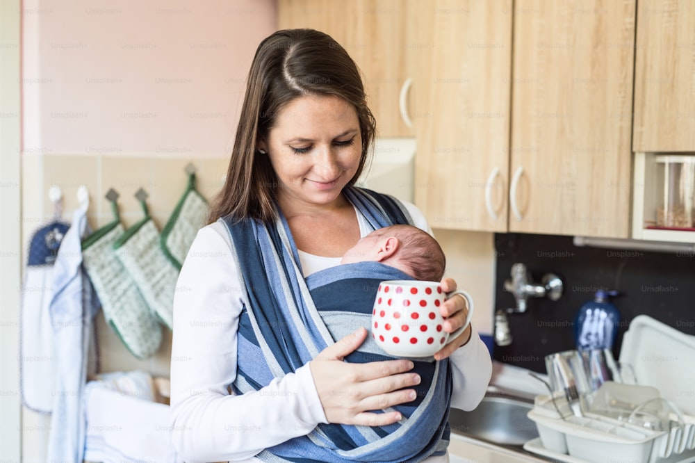 Schöne junge Mutter in der Küche mit ihrem neugeborenen Sohn im Tragetuch zu Hause, der eine rot gepunktete Tasse hält