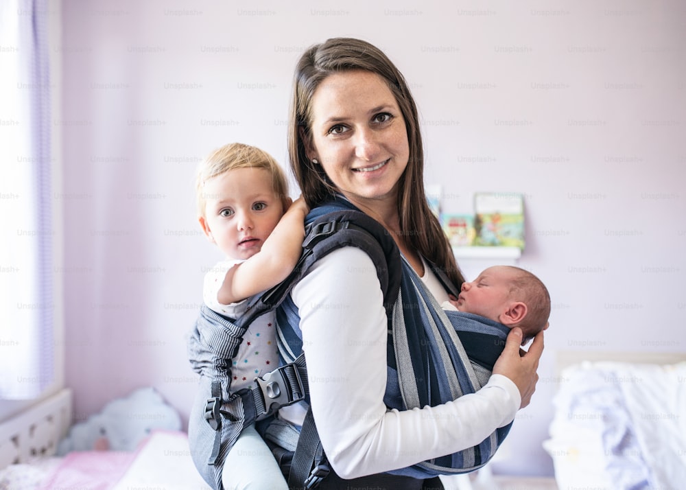 Hermosa madre joven con su hijo recién nacido en cabestrillo y su hija en portabebés en casa