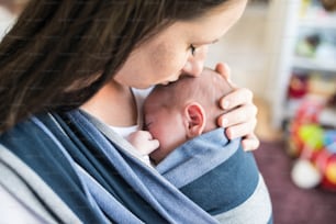 Gros plan d’une jeune mère méconnaissable avec son nouveau-né en écharpe à la maison, l’embrassant