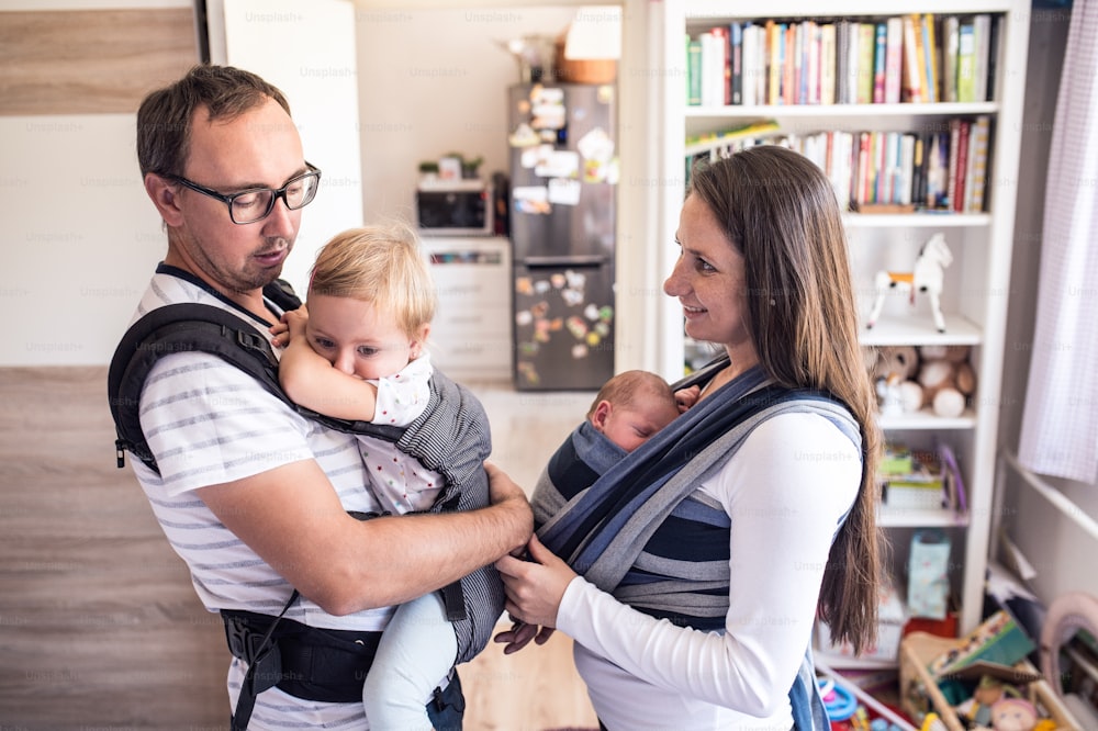 Beautiful young mother with her newborn son in sling and father with his daughter in baby carrier at home