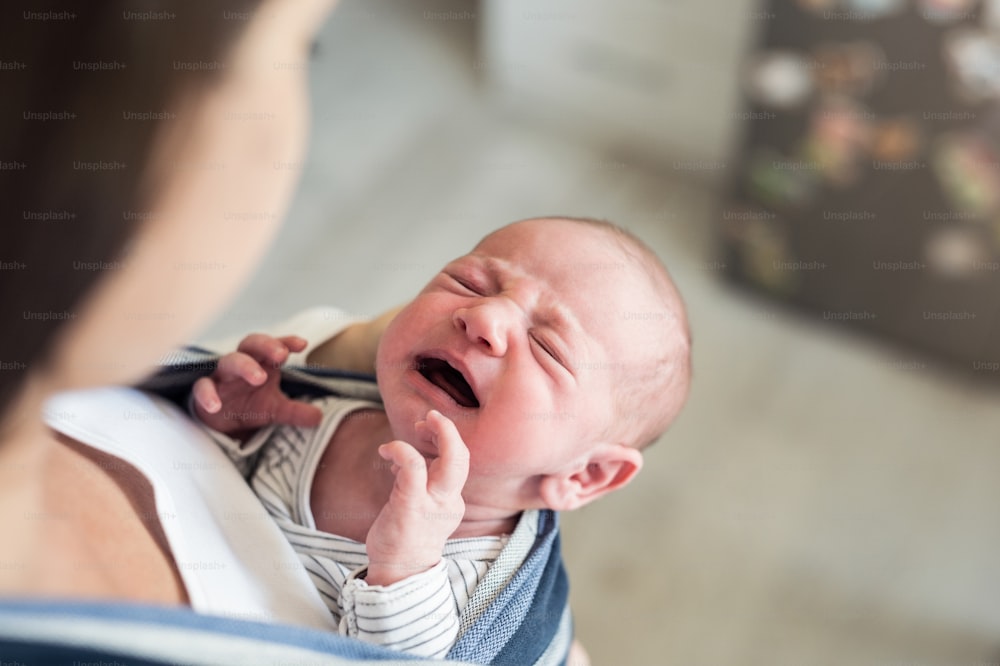 Primer plano de una joven madre irreconocible con su bebé recién nacido llorando en cabestrillo en casa