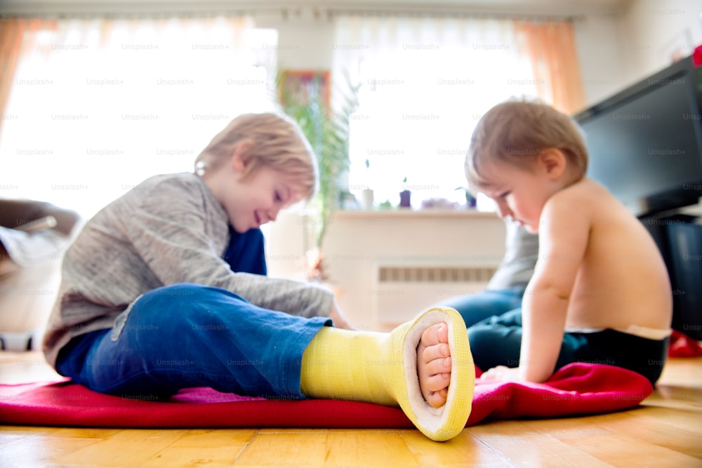 Cute little blond boy with broken leg in cast with his little brother sitting on the wooden floor, playing. Childrens daytime fun. Happy to be at home.