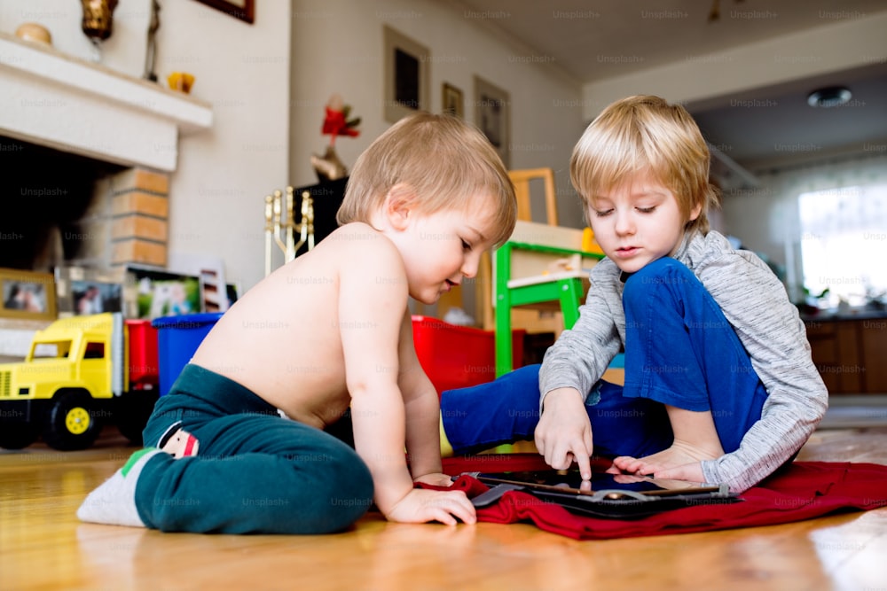 Cute little blond boy with broken leg in cast with his little brother sitting on the wooden floor, playing on tablet. Childrens daytime fun. Happy to be at home.