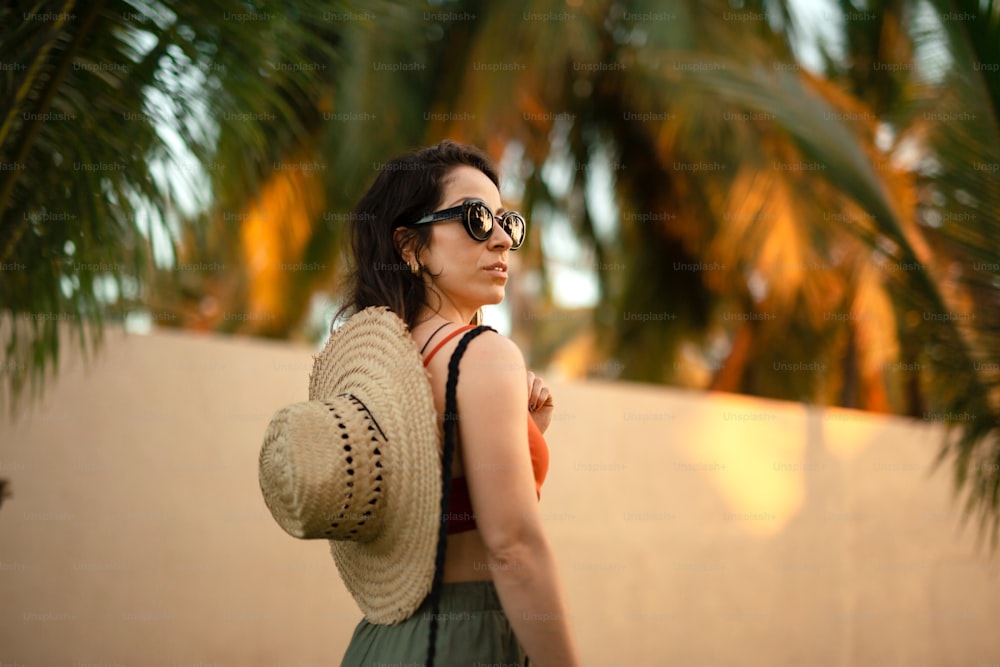 Une femme portant un chapeau et des lunettes de soleil se tient devant un palmier