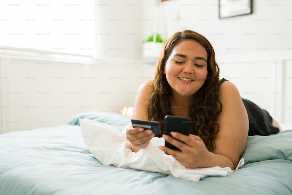 Aufgeregte junge Frau im Bett, die ihr Smartphone benutzt und mit einer Kreditkarte für ihren Online-Einkauf bezahlt