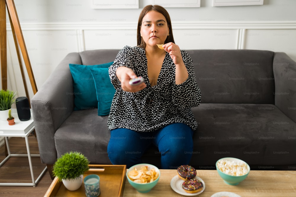 Alimentación poco saludable. Mujer joven con sobrepeso que usa el control remoto y mira películas mientras come comida chatarra