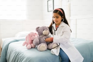 Cute latin elementary girl with a white lab coat and a stethoscope playing to be doctor with a teddy bear on her bedroom