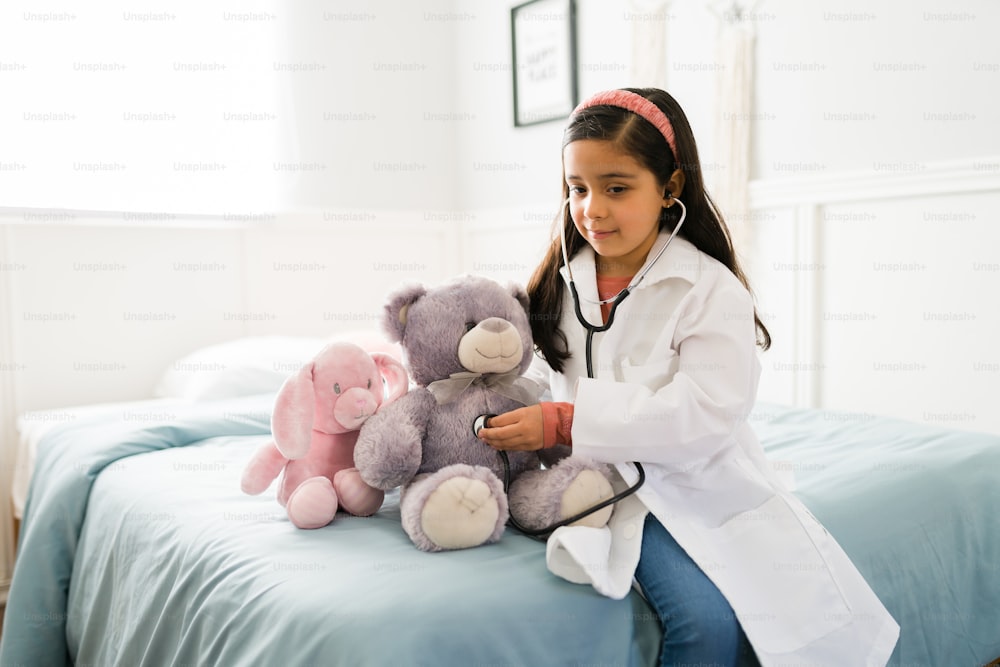 Linda niña latina de primaria con una bata blanca de laboratorio y un estetoscopio jugando a ser médico con un oso de peluche en su habitación
