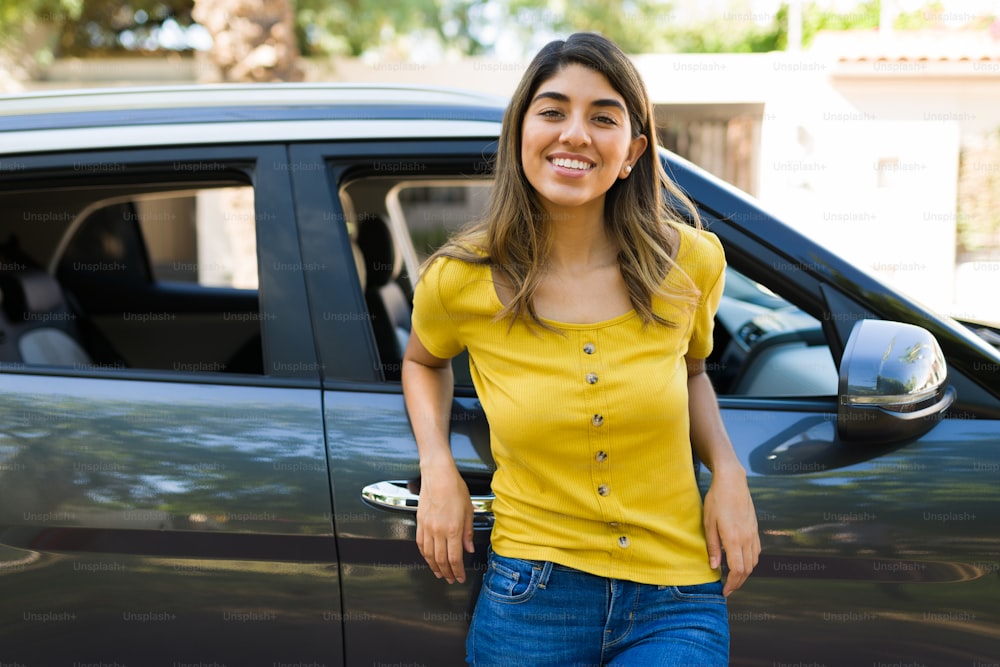 Mulher atraente sorrindo e fazendo contato visual enquanto está ao lado do carro preto fora de sua casa