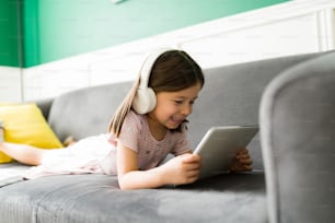 Excited elementary kid relaxing on the couch while watching online videos or movies on the tablet at home
