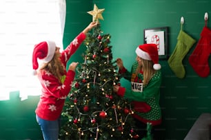 Mujeres jóvenes festivas con suéteres feos decorando su árbol de Navidad y poniendo la estrella en la parte superior