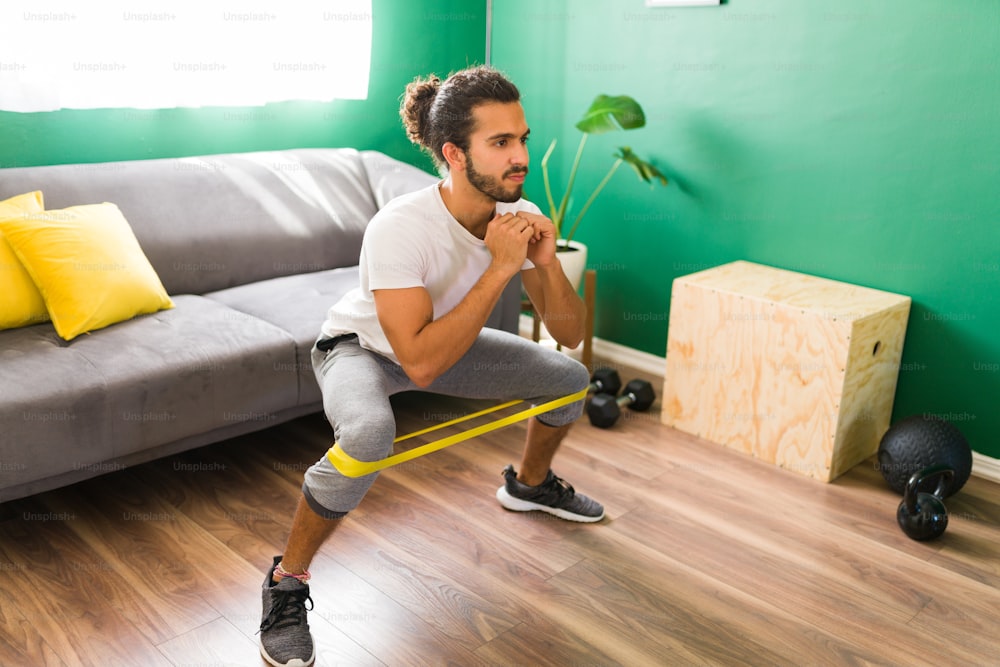 Attractive man in his 20s squatting with a fitness band on his legs. Fitness man exercising in the living room