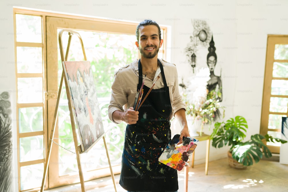 Atractivo joven sosteniendo sus pinceles y una paleta lista para crear una pintura artística en el estudio