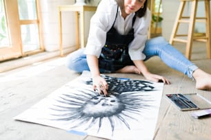 Close up de uma jovem mulher desenhando um retrato artístico a carvão enquanto sentada no chão do estúdio de arte
