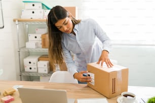 Hispanic young woman working at her startup business and putting a shipping label on a package with beauty products
