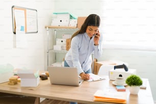 Cheerful female entrepreneur talking on the phone with a customer or her supplier and writing down information while working at her business startup