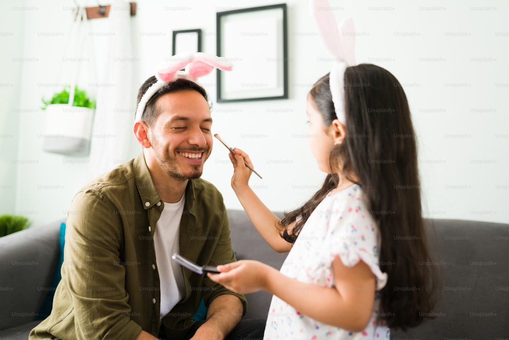 Cheerful dad with bunny ears playing with her little daughter. Beautiful girl putting makeup and eyeshadow to her father