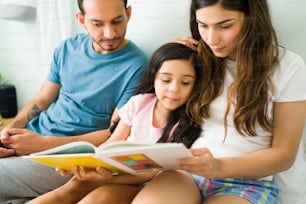 Family reading time. Hispanic mother and dad reading a children's book to her little daughter while resting in bed together