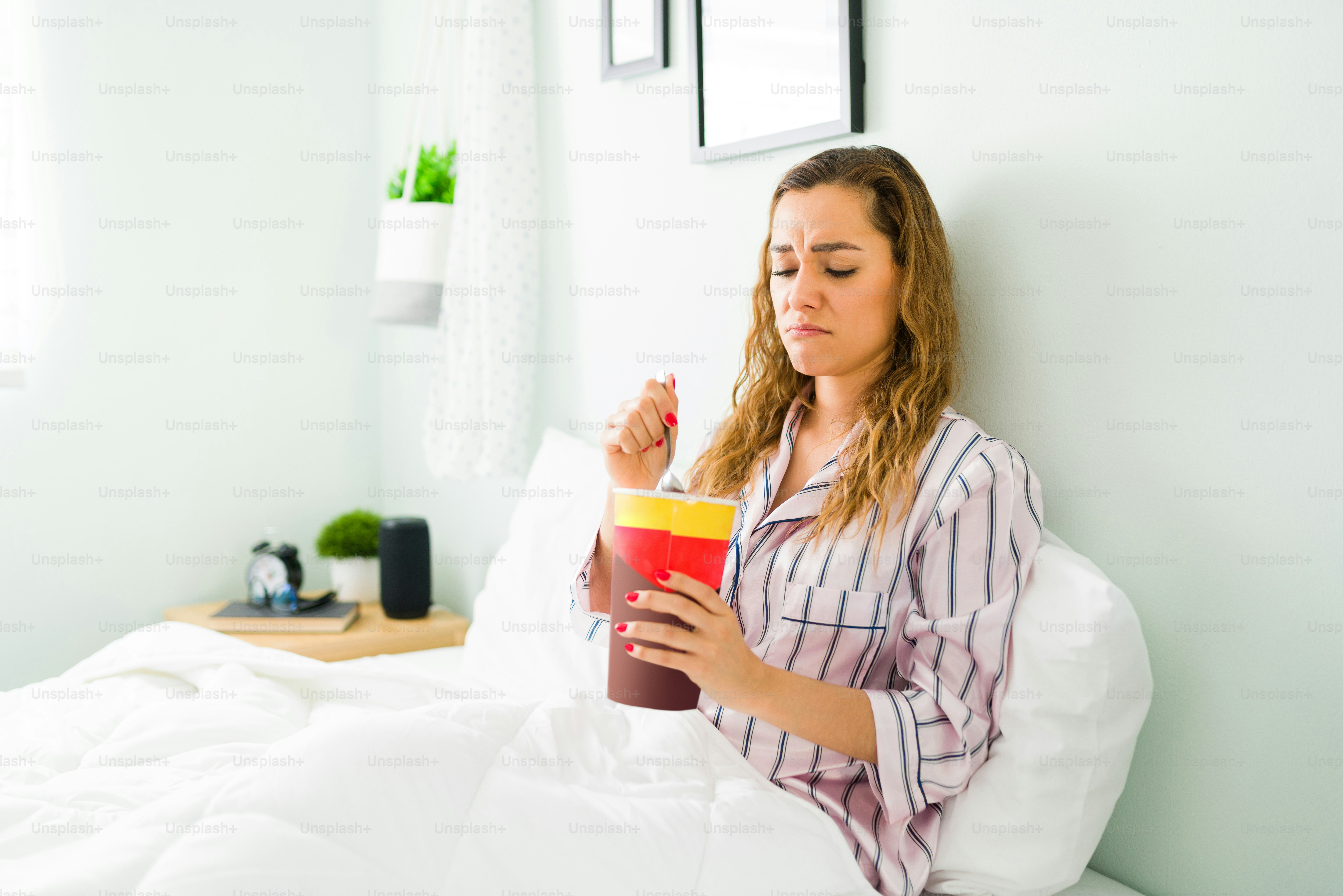 Depressed woman in her 30s feeling sad and heartbroken while resting in her pajamas. Hispanic woman eating chocolate ice cream in the bedroom