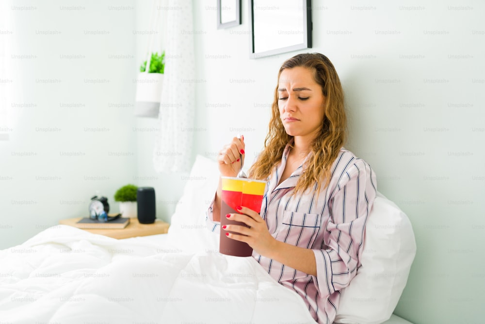 Mujer deprimida de unos 30 años que se siente triste y desconsolada mientras descansa en pijama. Mujer hispana comiendo helado de chocolate en el dormitorio