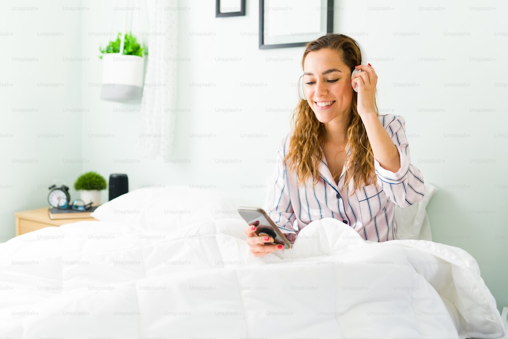 Aufgeregte lateinamerikanische Frau, die Musik mit Kopfhörern hört, während sie sich in ihrem sauberen weißen Bett ausruht. Glückliche Frau, die ein Lied mit ihrem Smartphone genießt