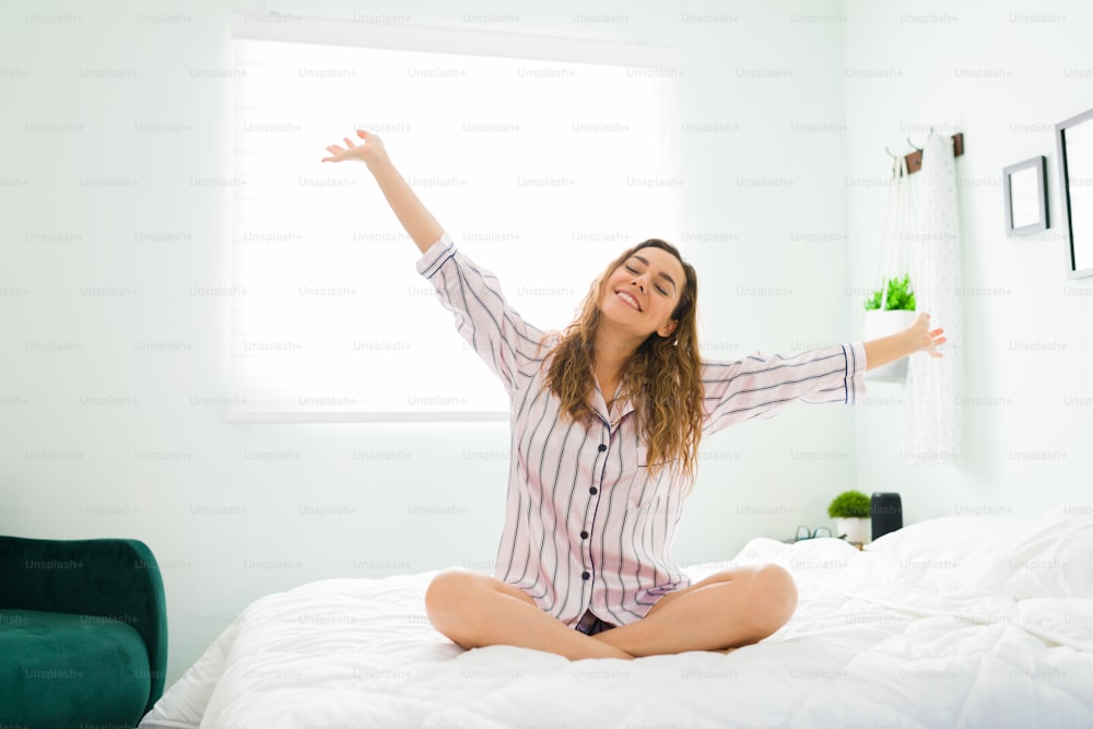 Attraktive positive Frau mit Pyjama, die nach dem Aufwachen im Bett sitzt und ihre Arme ausstreckt. Hispanische Frau fühlt sich glücklich und ausgeruht