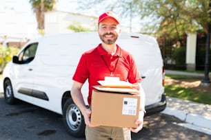 Retrato de un mensajero feliz con una furgoneta blanca sonriendo mientras sostiene algunos paquetes y paquetes antes de entregarlos fuera de la puerta de una casa