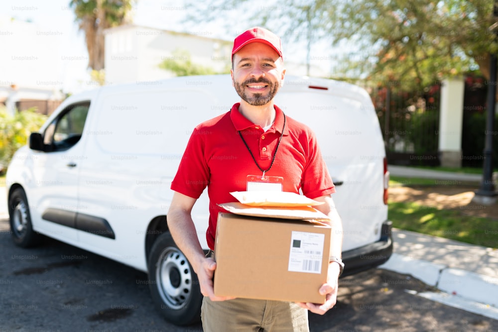 Porträt eines glücklichen Lieferkuriers mit einem weißen Lieferwagen, der lächelt, während er einige Pakete und Pakete hält, bevor er sie vor der Tür eines Hauses ausliefert