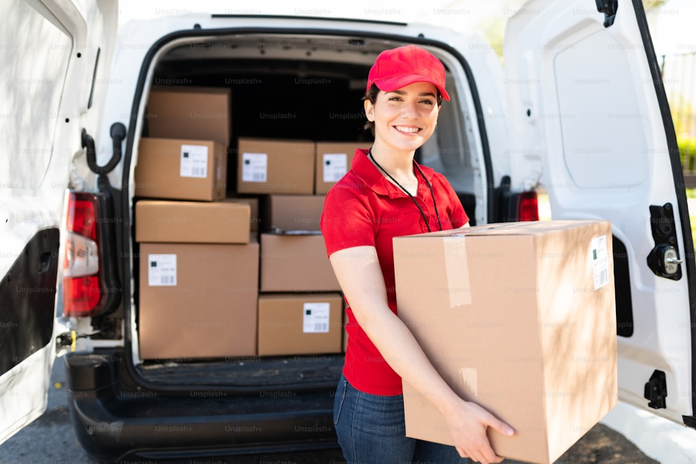 Porträt einer attraktiven kaukasischen Arbeiterin in roter Uniform, die lächelnd Pakete auslädt und neben einem Lieferwagen mit vielen Kisten und Paketen steht