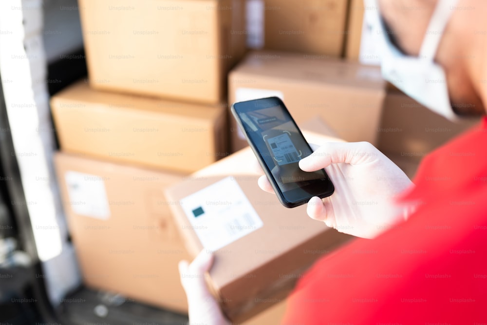 Close up of a male worker and delivery person with a face mask holding a smartphone and scanning the barcode of a shipping label to deliver a package