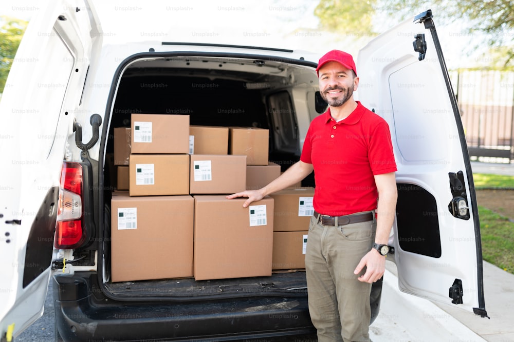 Happy delivery man standing in the back of a white truck with a lot of packages. Attractive man in his 30s in a red uniform working as a delivery courier