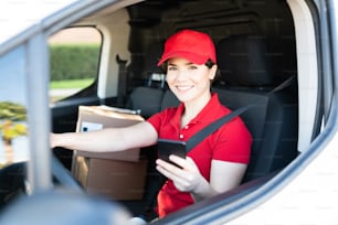 Retrato de uma bela mulher caucasiana com um uniforme vermelho sorrindo e segurando um smartphone enquanto está sentado ao volante de uma van de entrega com pacotes