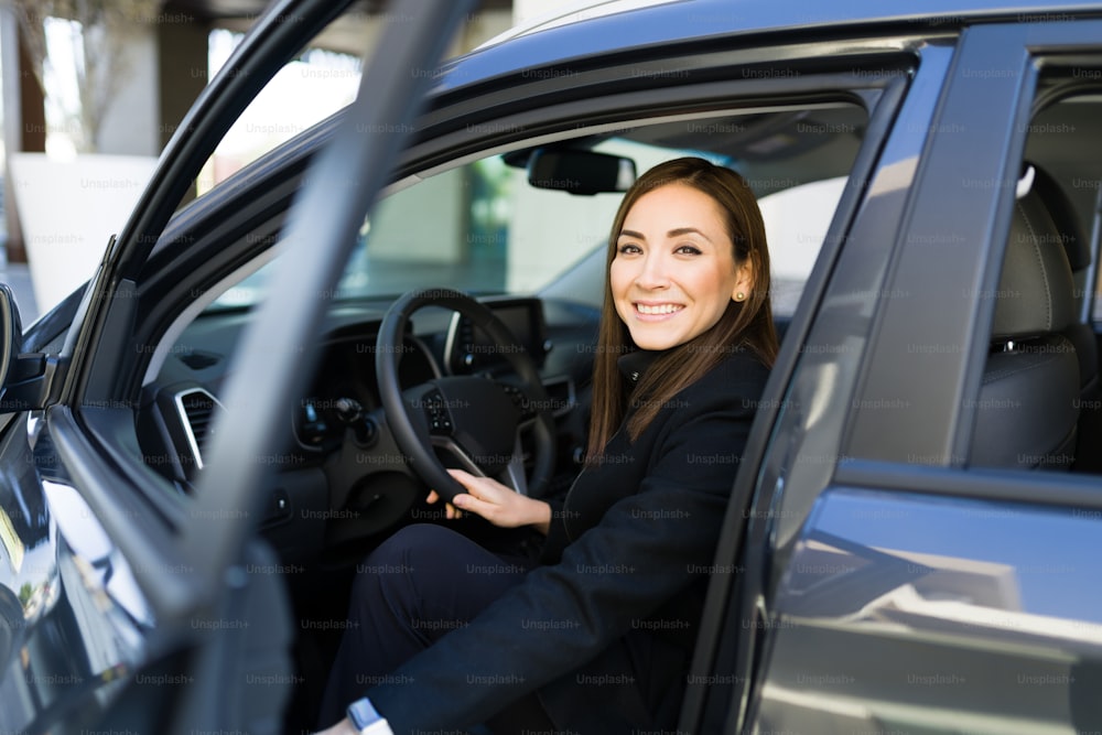 Mujer de negocios feliz haciendo contacto visual mientras sostiene la puerta del automóvil y se sienta en el asiento del conductor antes de ir a la oficina o cita de trabajo