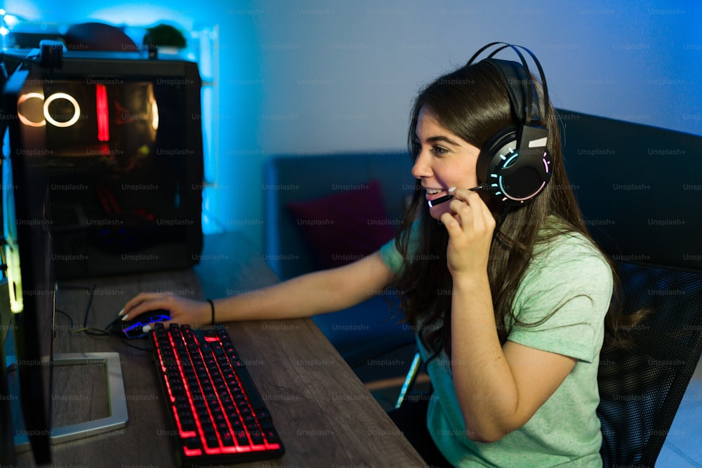 백인 젊은 여성이 LED 조명이 있는 게임용 컴퓨터에서 비디오 게임을 하는 동안 마이크에서 온라인 플레이어와 이야기합니다