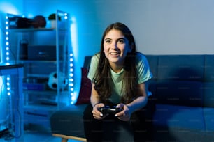 Joven jugadora divirtiéndose y usando un control remoto para jugar un videojuego mientras está sentada en el sofá de su habitación decorada con luces led