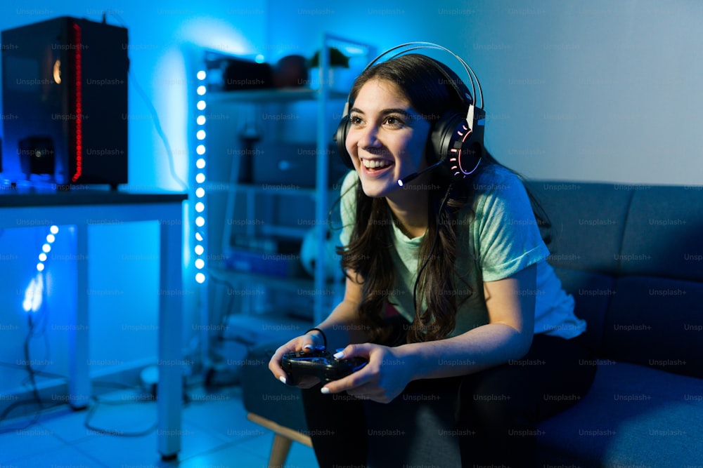 リモコンでビデオゲームで遊んで勝つときに笑顔の幸せな女性ゲーマー。寝室のソファに座ってネオンLEDライトを持つ若い女性
