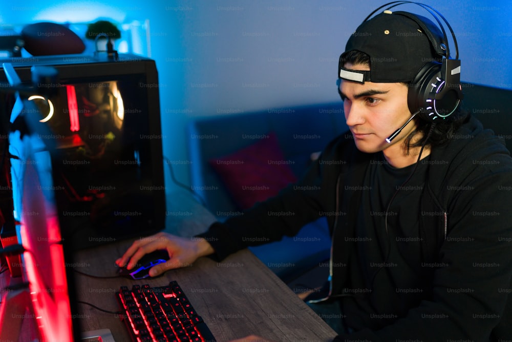 Jeune homme séduisant avec des écouteurs et un microphone regardant l’écran d’un ordinateur de jeu PC et jouant à un jeu vidéo en ligne