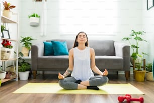Calm and relaxed latin woman in an easy yoga pose with her eyes closed and doing a meditation routine in the living room floor