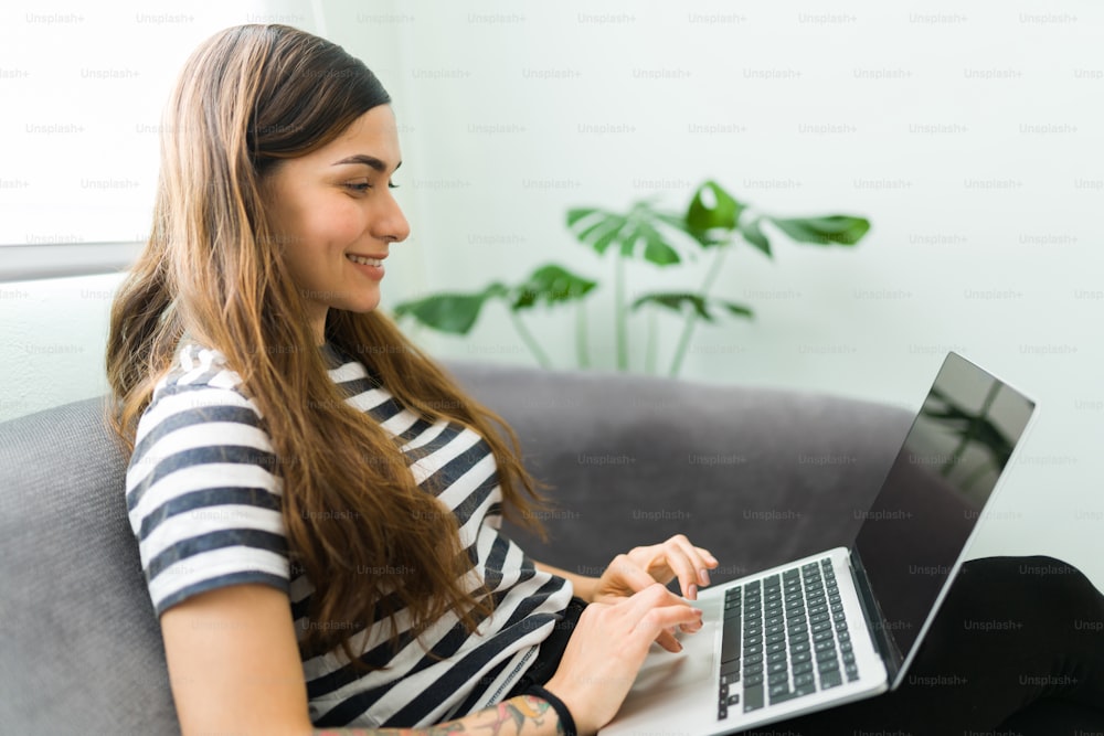 Junge Frau lächelt, während sie in ihrem Laptop online einkauft. Glückliche Frau in ihren 20ern, die sich soziale Medien anschaut und mit ihren Online-Freunden chattet