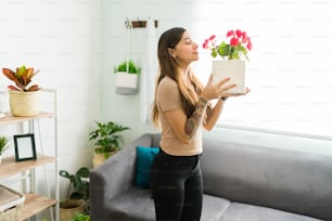 Mujer relajada y tranquila oliendo una flor en una maceta mientras está de pie en su sala de estar. Hermosa joven con una salud mental positiva